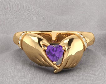 May include: A gold-colored ring featuring two hands cradling a heart-shaped purple gemstone. The ring has small, clear stones set on each side of the band.