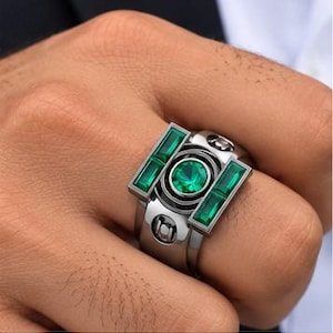 May include: A silver-toned metal ring featuring a central round green gemstone flanked by two rectangular green baguette-cut gemstones. The band has a polished finish with circular accents and a structured, geometric design.