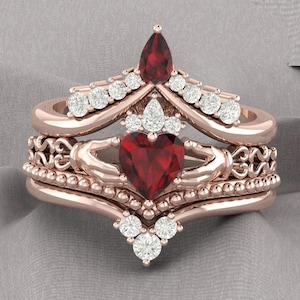 May include: A three-piece rose gold engagement ring set with a red heart-shaped center stone and a pear-shaped red stone on the top band. The ring is adorned with smaller clear stones.