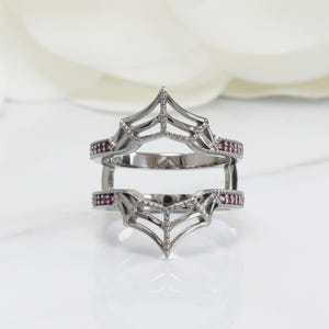 Peut inclure: Une protection de bague argentée au design unique. La protection de bague présente un motif en forme de toile d'araignée avec de petites pierres précieuses rouges rondes serties sur les bords. La protection de bague est conçue pour être portée avec une autre bague.