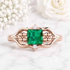 Peut inclure: Bague en or rose ornée d'une émeraude carrée au centre et de deux petits diamants. La bague présente un design orné avec des détails en forme de chaîne sur les côtés. Posée sur une surface en marbre blanc.