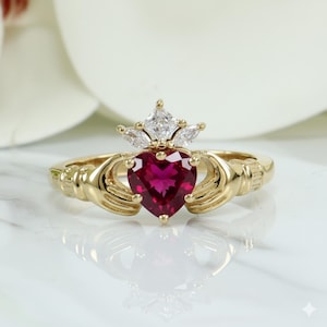Könnte beinhalten: Ein goldfarbener Ring mit einem herzförmigen, tiefroten Edelstein und einer Krone aus klaren Steinen. Das Design umfasst ineinandergreifende Hände auf beiden Seiten des Herzens, mit aufwendigen Details und poliertem Finish.