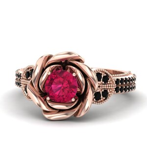 May include: Rose gold engagement ring featuring a central, round, ruby-colored gemstone. The ring's design incorporates a rose-shaped setting and is accented with black gemstones along the band, creating a romantic and elegant aesthetic.