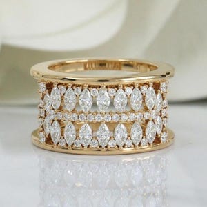 May include: A gold-toned ring with multiple rows of sparkling, clear gemstones. The ring features a band with a series of marquise-cut stones and smaller round stones, creating a luxurious and elegant design. The ring is set against a soft, blurred background.