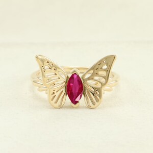 May include: A gold-colored ring featuring a butterfly design. The butterfly wings are intricately detailed, and a marquise-cut, ruby-colored gemstone is set in the center. The ring is a delicate piece of jewelry.