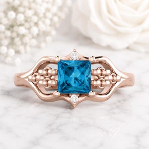 Op de afbeelding: Roségouden ring met een vierkante, helderblauwe edelsteen. De ring heeft een decoratief ontwerp met kleine diamantaccenten. De band heeft een opengewerkt patroon. De ring staat tegen een zachte, vage achtergrond.