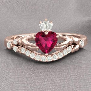 Peut inclure: Une bague en or rose avec une pierre précieuse en rubis en forme de cœur entourée de petits diamants. La bague présente un motif de nœud celtique avec deux mains jointes.