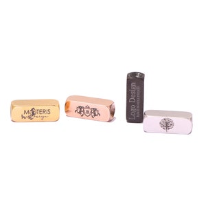 Pode incluir: Três pingentes de barra de metal diferentes com designs gravados. Os pingentes são dourados, ouro rosa e preto. O pingente dourado tem o texto "MOTERIS Parfume" gravado. O pingente de ouro rosa tem um brasão com a letra "B" gravada. O pingente preto tem o texto "Logo Design Made in Hollywood" gravado.