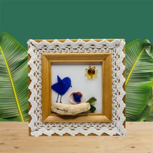 May include: A framed artwork featuring a blue glass bird perched on driftwood. The artwork includes a small sunflower and a seashell with blue glass accents. The frame is wood with a white lace border, set against a green background.