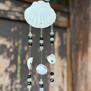 May include: A hanging mobile featuring a large white scallop shell at the top, suspended by a gold chain. Three vertical strands hang from the shell, decorated with alternating clear faceted beads, dark green beads, and small gold-toned spacers.