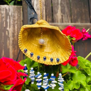May include: A decorative wind chime made from a gold-painted seashell, with blue beads and a crystal ball. Strands of white, blue, and shell beads hang below. The chime is suspended by a gold chain. The background includes red flowers and green foliage.