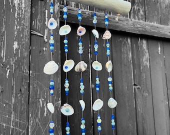 Puede incluir: Un móvil colgante que presenta una pieza horizontal de madera flotante de color claro con cinco hilos verticales suspendidos. Cada hilo está decorado con una variedad de cuentas de vidrio azul y conchas blancas de diversas formas y tamaños.