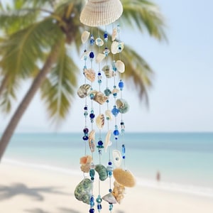 May include: A seashell wind chime featuring a large, ridged shell at the top, with strands of smaller shells and blue beads. The chime hangs against a blurred beach scene with palm trees and a blue sky.