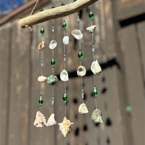 May include: A handcrafted wind chime featuring a natural driftwood branch with hanging strands of seashells, green glass beads, and clear crystal beads. The seashells vary in size and shape, creating a unique coastal decor piece.