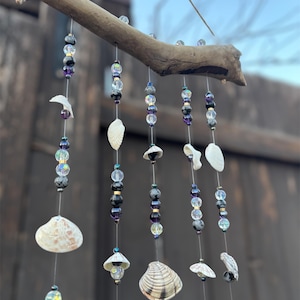 May include: A handmade wind chime with a natural wooden branch, strands of beads, and seashells. The beads are iridescent, black, and purple. The seashells are white and beige. The wind chime is suspended outdoors.