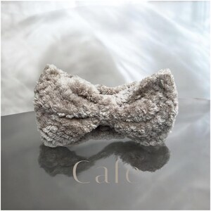 K&ouml;nnte beinhalten: Ein flauschiges, hellbraunes Stirnband mit Schleifen-Design. Das Material hat eine strukturierte Oberfl&auml;che, die das Licht einf&auml;ngt. Das Wort "Cafe" ist im unteren Teil des Bildes zu sehen.