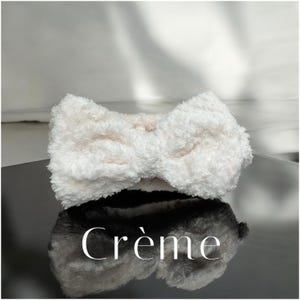 K&ouml;nnte beinhalten: Ein flauschiges, cremefarbenes Stirnband mit Schleifendesign. Das Stirnband besteht aus einem weichen, strukturierten Material. Das Wort "Cr&egrave;me" wird in wei&szlig;er, eleganter Schrift angezeigt.