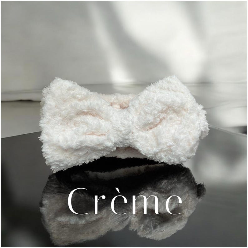 K&ouml;nnte beinhalten: Ein flauschiges, cremefarbenes Stirnband mit Schleifendesign. Das Stirnband besteht aus einem weichen, strukturierten Material. Das Wort "Cr&egrave;me" wird in wei&szlig;er, eleganter Schrift angezeigt.