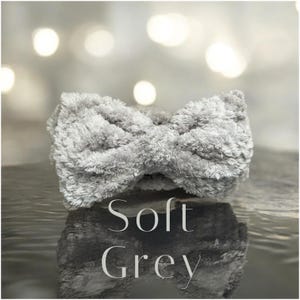 K&ouml;nnte beinhalten: Ein weiches, hellgraues Stirnband mit Schleifendesign. Das Stirnband besteht aus einem pl&uuml;schigen, strukturierten Material. Die Worte "Soft Grey" stehen in wei&szlig;er Schrift unter dem Stirnband.