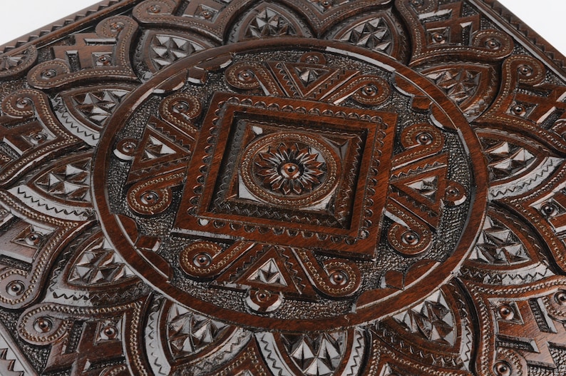 May include: A dark brown wooden box with intricate carved designs. The box has a square shape and a raised, circular design in the center.