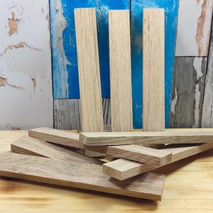 Könnte beinhalten: Eine Sammlung unfertiger Holzbretter und -leisten in hellem Farbton. Einige Bretter stehen aufrecht, andere sind horizontal angeordnet. Die Holzmaserung ist sichtbar, und die Teile variieren in Größe und Form, was darauf hindeutet, dass sie für ein Bastel- oder Holzbearbeitungsprojekt bestimmt sind.