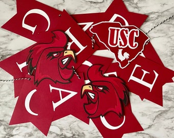 May include: Red and white banner with the letters "GAME" and a "USC" logo. The banner features a red rooster mascot and is strung with black and white twine. The banner is a festive decoration.