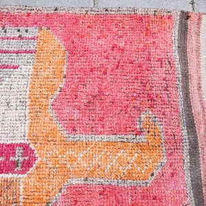 May include: Close-up of a pink and orange woven rug with a geometric pattern. The rug has a fringe border and is made of a soft, textured material.