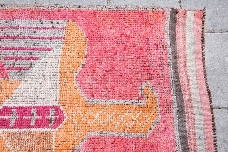 May include: Close-up of a pink and orange woven rug with a geometric pattern. The rug has a fringe border and is made of a soft, textured material.