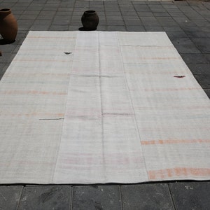 K&ouml;nnte beinhalten: Ein gro&szlig;er, rechteckiger, cremefarbener Teppich mit Patchwork-Design. Der Teppich hat dezente orange und rote Streifen und kleine dekorative Elemente. Der Teppich liegt auf einer dunkelgrauen Fliesenoberfl&auml;che, mit einer Holzbank und T&ouml;pferwaren im Hintergrund.