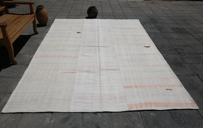 K&ouml;nnte beinhalten: Ein gro&szlig;er, rechteckiger, cremefarbener Teppich mit Patchwork-Design. Der Teppich hat dezente orange und rote Streifen und kleine dekorative Elemente. Der Teppich liegt auf einer dunkelgrauen Fliesenoberfl&auml;che, mit einer Holzbank und T&ouml;pferwaren im Hintergrund.