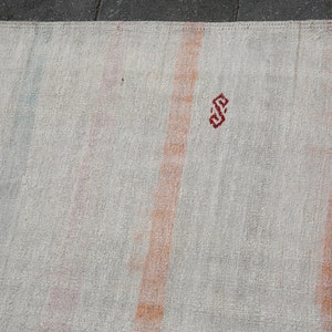 K&ouml;nnte beinhalten: Cremefarbener Teppich mit vertikalen Streifen in Orange-, Rosa- und Blaut&ouml;nen. Oben ist ein kleines, rotes geometrisches Design zu sehen. Der Teppich hat ein strukturiertes Aussehen, was auf eine gewebte oder handgefertigte Konstruktion hindeutet.