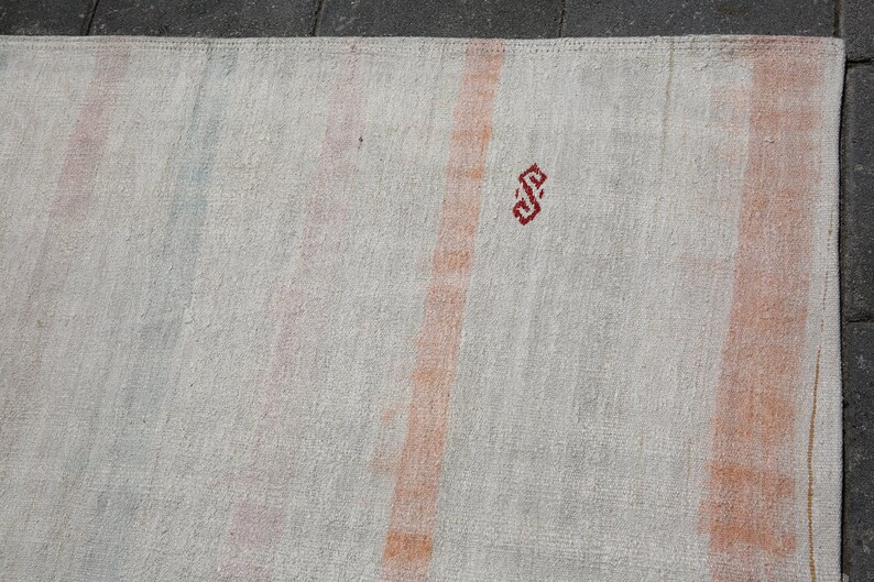 K&ouml;nnte beinhalten: Cremefarbener Teppich mit vertikalen Streifen in Orange-, Rosa- und Blaut&ouml;nen. Oben ist ein kleines, rotes geometrisches Design zu sehen. Der Teppich hat ein strukturiertes Aussehen, was auf eine gewebte oder handgefertigte Konstruktion hindeutet.