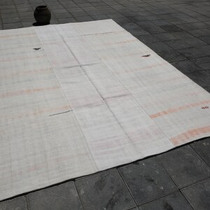 K&ouml;nnte beinhalten: Ein gro&szlig;er, rechteckiger, cremefarbener Teppich mit verblassten orangefarbenen Streifen und Flecken. Der Teppich liegt auf einer dunkelgrauen, ziegelartigen Oberfl&auml;che. Auf der Oberfl&auml;che des Teppichs sind einige kleine, dunkle Flecken zu sehen.