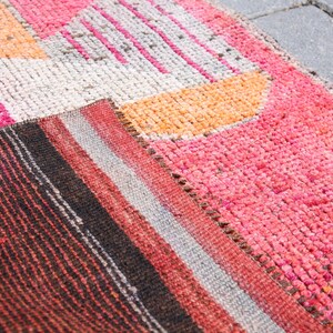 May include: A close-up of a colorful woven rug with geometric patterns in shades of pink, orange, white, and black. The rug is made of a soft, textured material and has a hand-woven look.