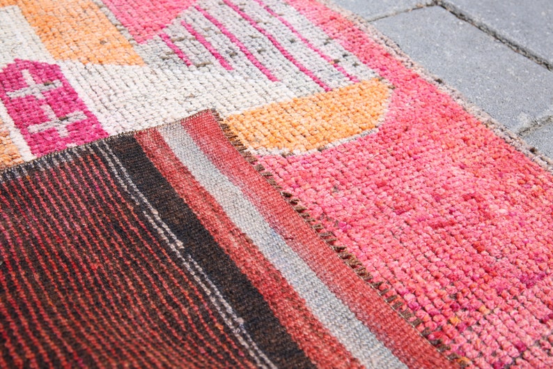 May include: A close-up of a colorful woven rug with geometric patterns in shades of pink, orange, white, and black. The rug is made of a soft, textured material and has a hand-woven look.