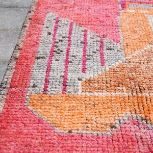 May include: Close-up of a pink, orange, and gray patterned rug with a geometric design. The rug is made of woven fabric and has a slightly distressed look.
