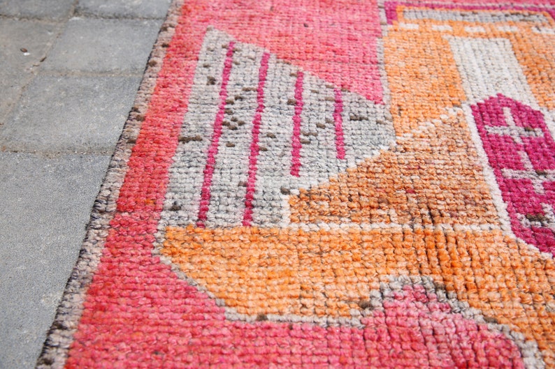 May include: Close-up of a pink, orange, and gray patterned rug with a geometric design. The rug is made of woven fabric and has a slightly distressed look.