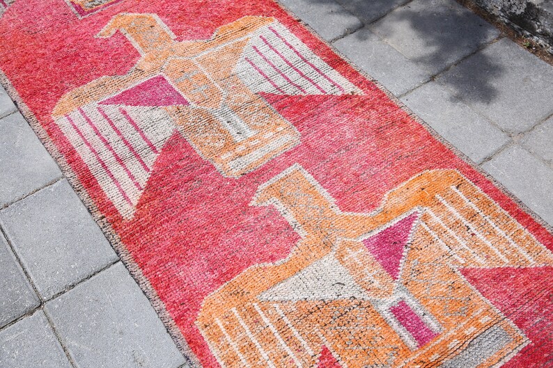 May include: A red, orange, and white patterned rug with a geometric design featuring stylized birds. The rug is made of woven fabric and has a distressed, vintage look.