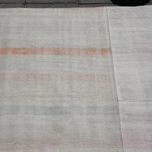K&ouml;nnte beinhalten: Ein rechteckiger, hellfarbener Teppich mit verblasstem, strukturiertem Aussehen. Der Teppich hat ein auff&auml;lliges horizontales Band in Orange-Rot sowie dezente rosa und blaue Streifen. Der Teppich besteht aus gewebtem Material.