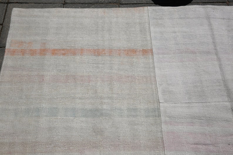K&ouml;nnte beinhalten: Ein rechteckiger, hellfarbener Teppich mit verblasstem, strukturiertem Aussehen. Der Teppich hat ein auff&auml;lliges horizontales Band in Orange-Rot sowie dezente rosa und blaue Streifen. Der Teppich besteht aus gewebtem Material.