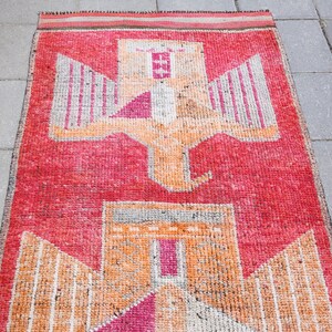 May include: A vintage red, orange, and white rug with a geometric pattern of two stylized birds. The rug has a fringed edge.