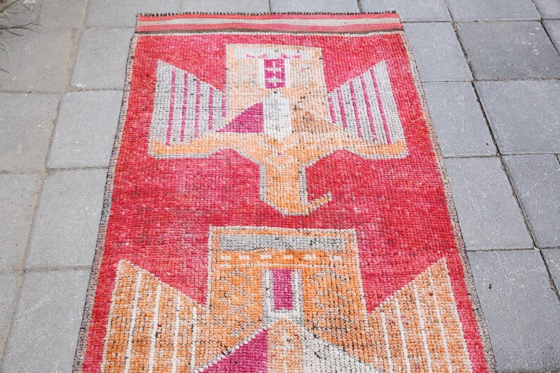May include: A vintage red, orange, and white rug with a geometric pattern of two stylized birds. The rug has a fringed edge.