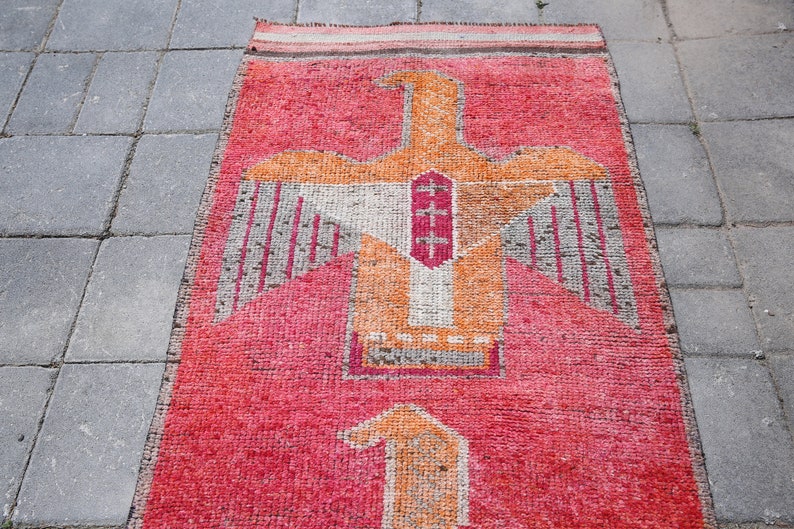 May include: A red, orange, and white rug with a stylized bird design. The rug has a fringed edge and is made of woven fabric.