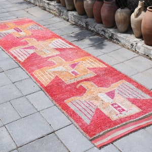 May include: A long, red runner rug with a geometric pattern of stylized birds in shades of orange, pink, and white. The rug is laid out on a gray concrete patio.