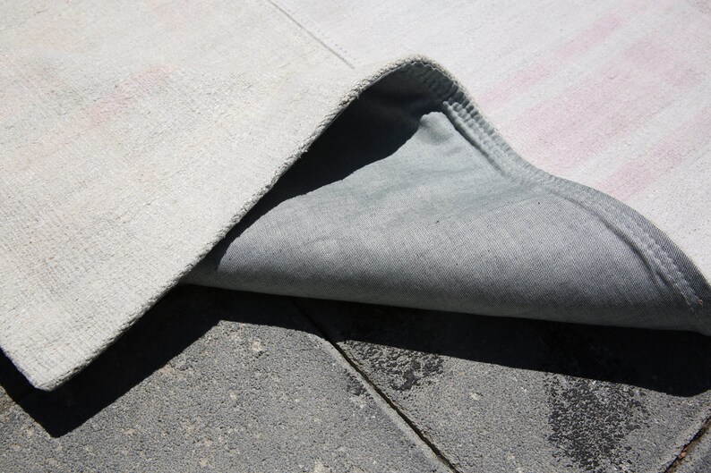 K&ouml;nnte beinhalten: Nahaufnahme eines wei&szlig;en Teppichs mit dezenten rosa Streifen, teilweise gefaltet, um eine graue R&uuml;ckseite zu zeigen. Die Textur des Teppichs ist sichtbar, auf einer strukturierten grauen Oberfl&auml;che.
