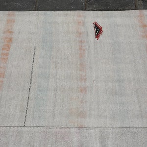 K&ouml;nnte beinhalten: Ein rechteckiger, cremefarbener Teppich mit vertikalen Streifen in Orange und Blau. Eine schwarze Linie verl&auml;uft diagonal &uuml;ber den Teppich. Ein kleines, dekoratives Element mit roten, schwarzen und wei&szlig;en geometrischen Mustern ist sichtbar.