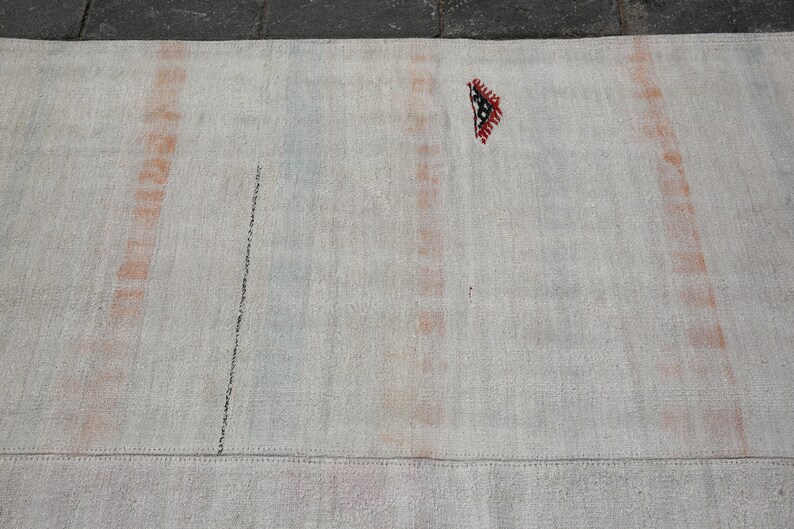 K&ouml;nnte beinhalten: Ein rechteckiger, cremefarbener Teppich mit vertikalen Streifen in Orange und Blau. Eine schwarze Linie verl&auml;uft diagonal &uuml;ber den Teppich. Ein kleines, dekoratives Element mit roten, schwarzen und wei&szlig;en geometrischen Mustern ist sichtbar.