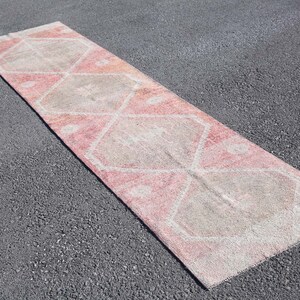 May include: A rectangular, distressed runner rug with a geometric pattern. The rug features a faded pink and beige color palette. The design includes diamond and floral shapes. The rug is placed on a dark gray surface.