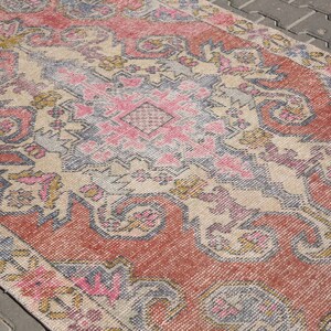 K&ouml;nnte beinhalten: Ein Vintage-Teppich mit einem geometrischen Muster in Rosa, Beige und Grau. Der Teppich hat einen abgenutzten Look und ist aus einem weichen, gewebten Material gefertigt.