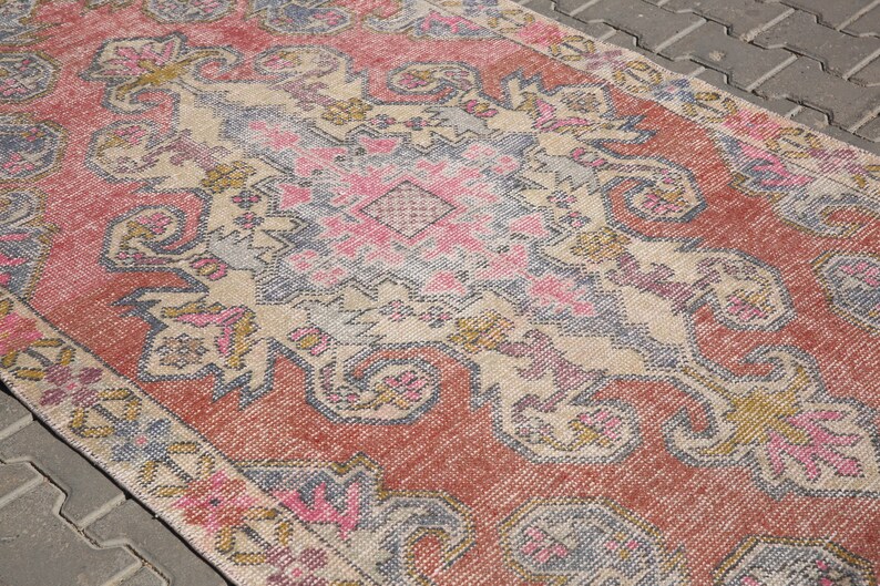 K&ouml;nnte beinhalten: Ein Vintage-Teppich mit einem geometrischen Muster in Rosa, Beige und Grau. Der Teppich hat einen abgenutzten Look und ist aus einem weichen, gewebten Material gefertigt.